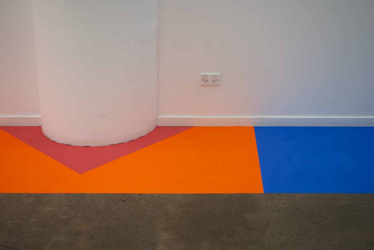 A pillar with a red powder paint triangle at its base sits inside an orange powder paint rectangle. A blue powder paint rectangle is to the right adjacent with the orange.